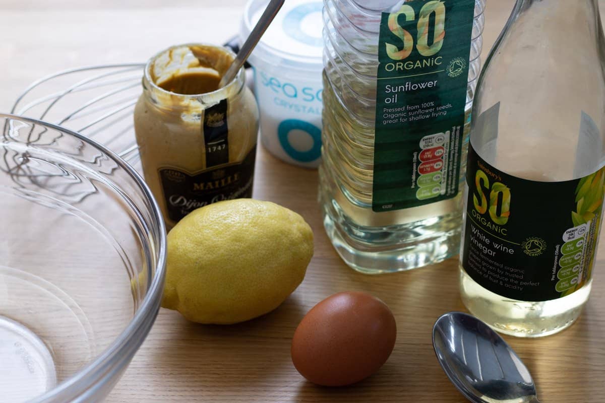 Lemon, oil, vinegar, salt, mustard and egg set out with glass bowl and spoon.