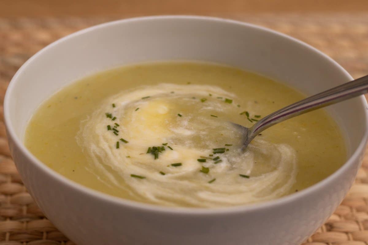 Leek and potato soup, with cream and chives, in a white bowl, on a raffia mat. A spoon is in the bowl.