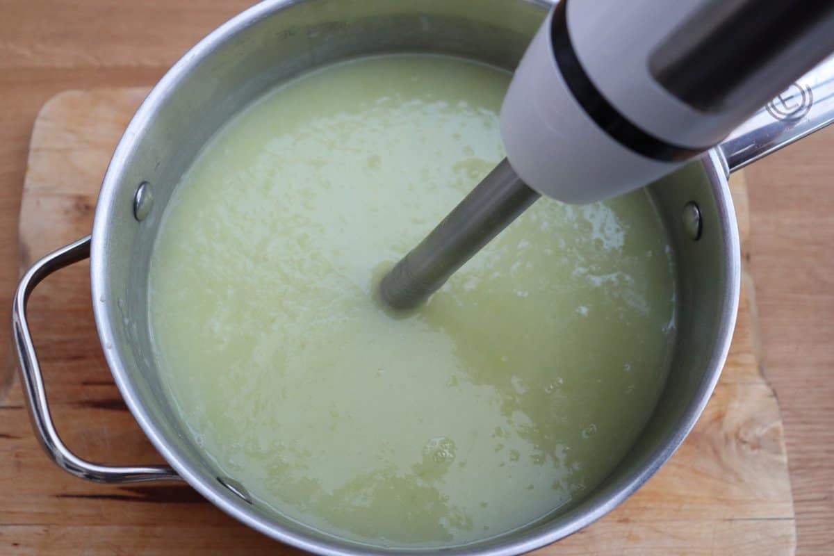 Leek and potato soup in a saucepan being blended by a hand blender.
