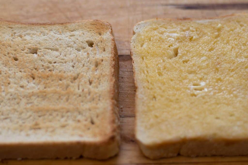 slice of toast toasted side up next to slice of buttered toast butter side up both on a wooden board