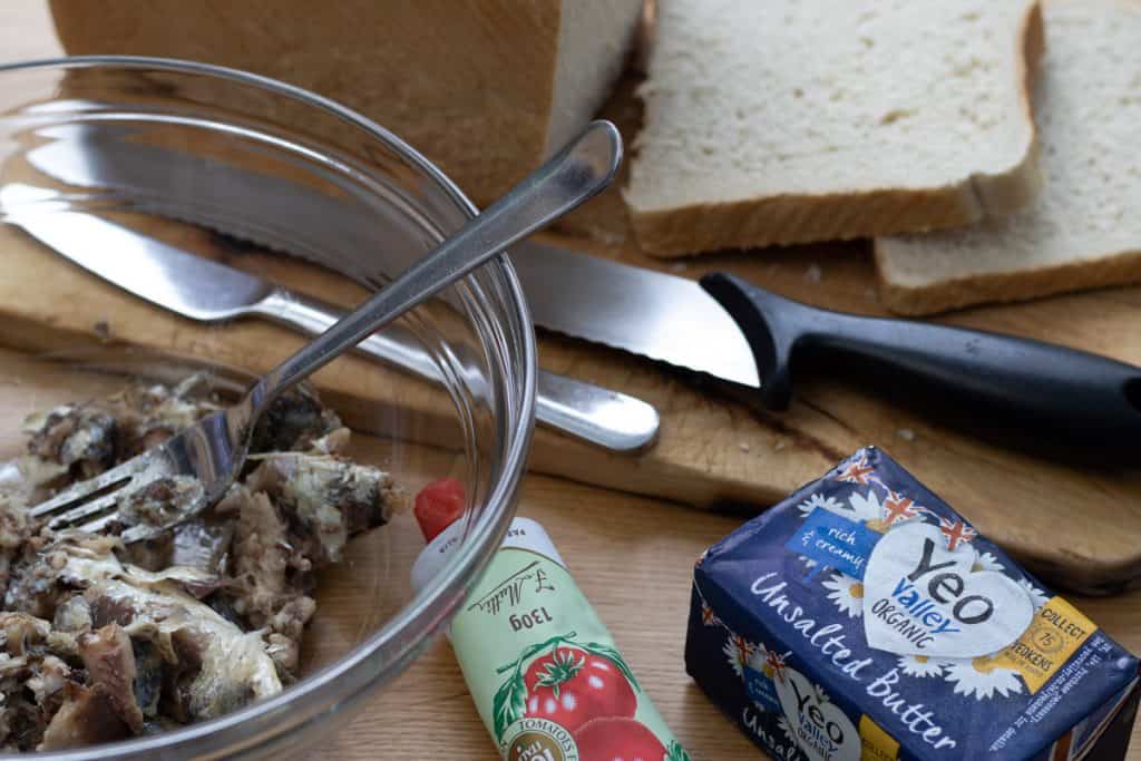 Ingredients for sardines on toast set out, sardines in a bowl, sliced bread on a wooden board, a tube of tomato puree and a pat of unsalted butter.