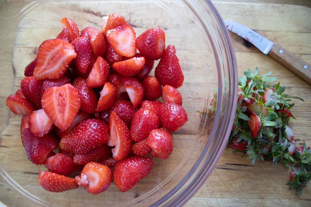 Hulled strawberries in a glass bowl next to the removed tops and a small knife.