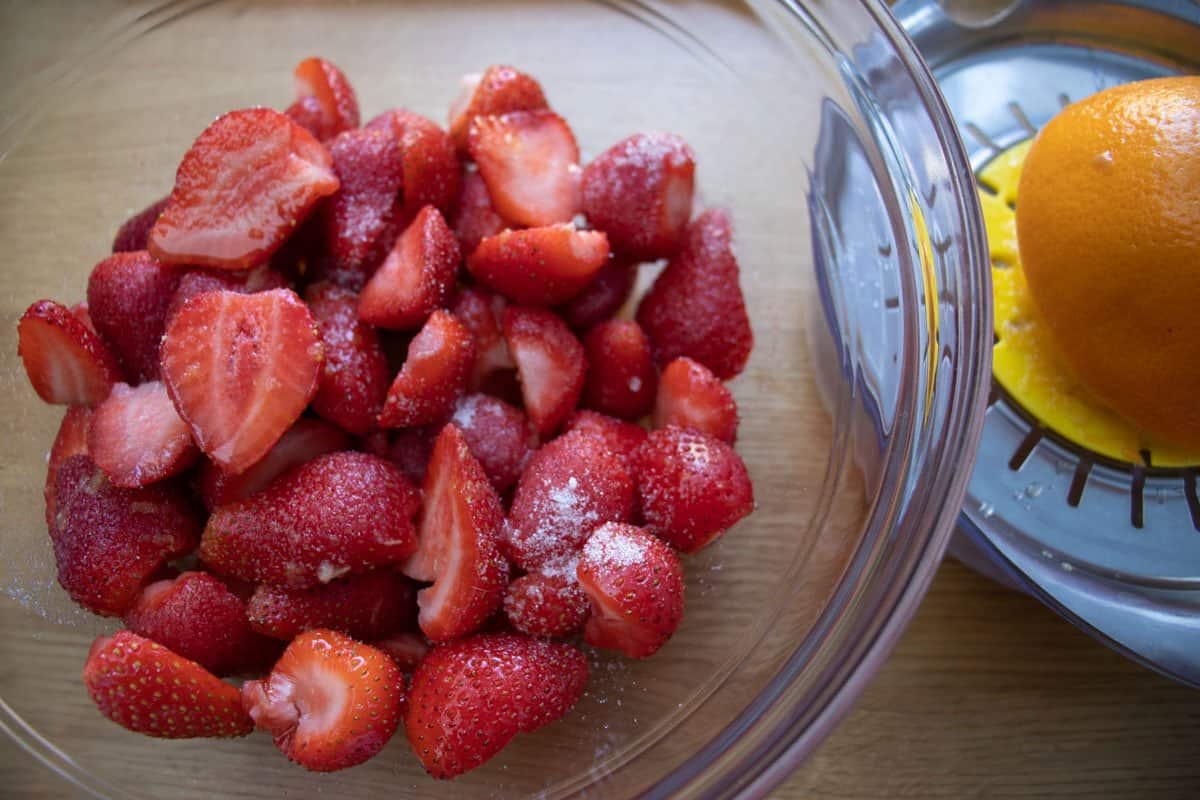Strawberries dusted with sugar in a glass bowl next to orange half on a hand juicer.