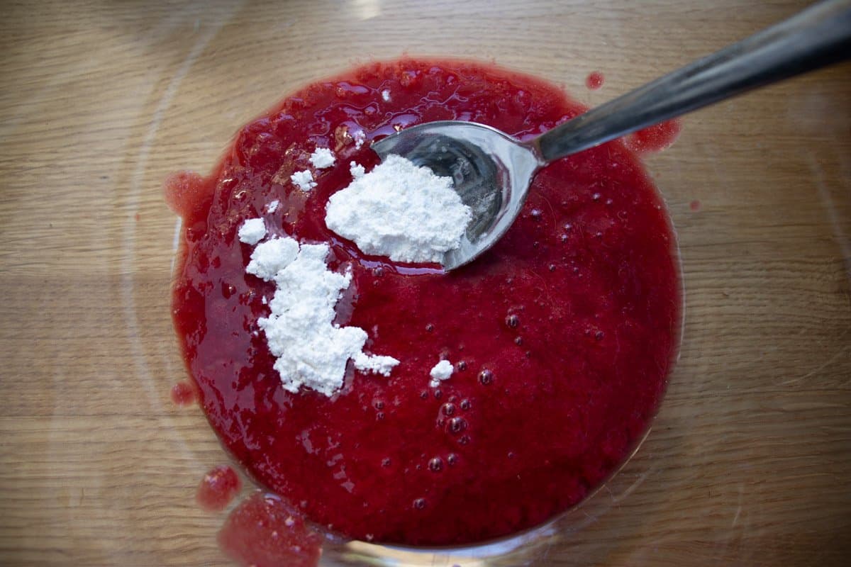 Raspberry coulis in a glass bowl with spoonful of icing sugar being added.
