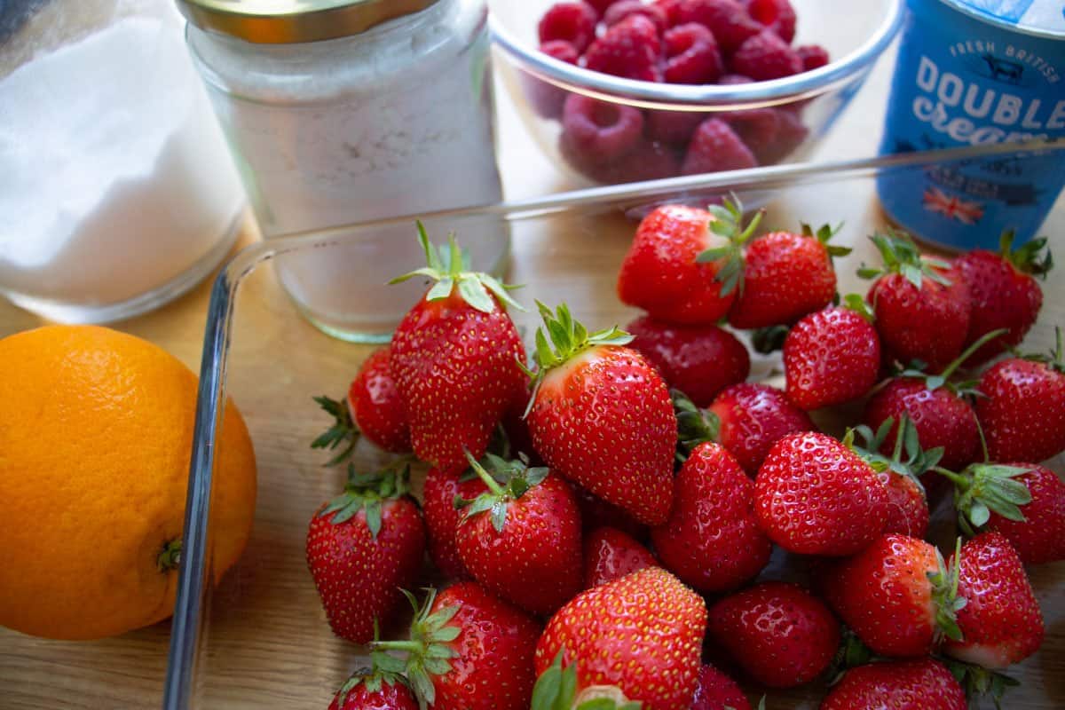 Strawberries in a glass dish with an orange, raspberries in a bowl, cream in a pot, sugar in a jar.
