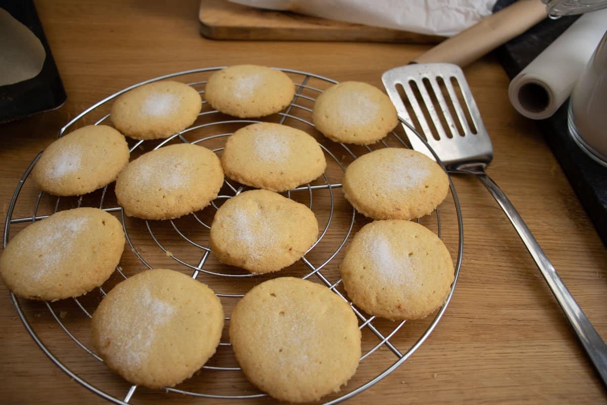 Baked lemon butter biscuits with caster sugar sprinkles on wire cooling rack with metal slice and rolls of greaseproof paper