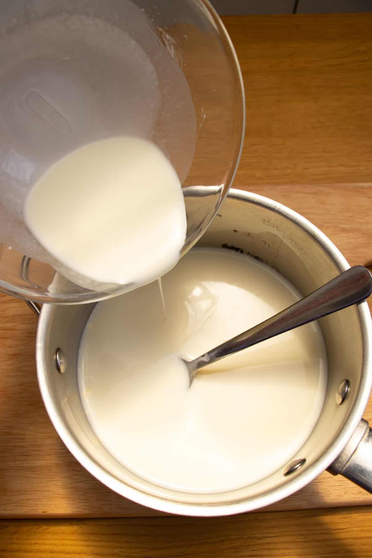 Cornflour (cornstarch) mixture being poured from a glass mixing bowl into a saucepan of milk.