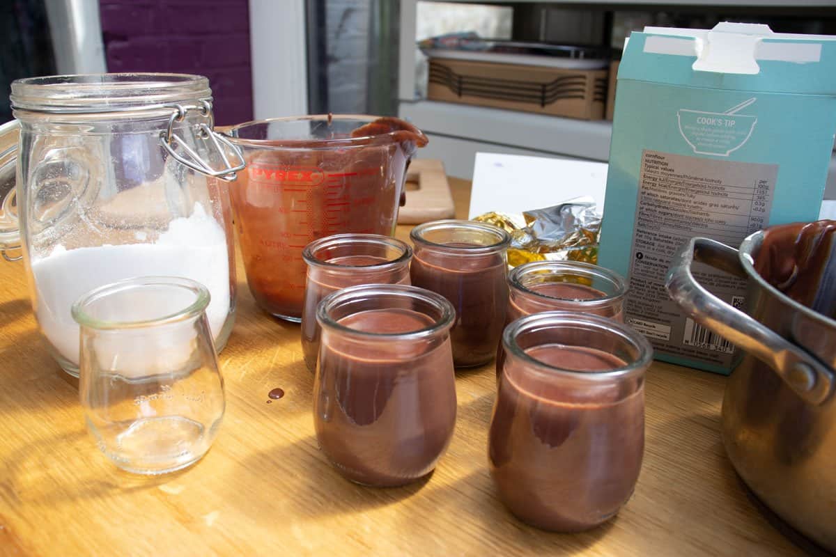 Glass jars filled with Dark Chocolate Cremeux on wooden surface with jar of sugar, and glass jug and saucepan both with some chocolate on them.