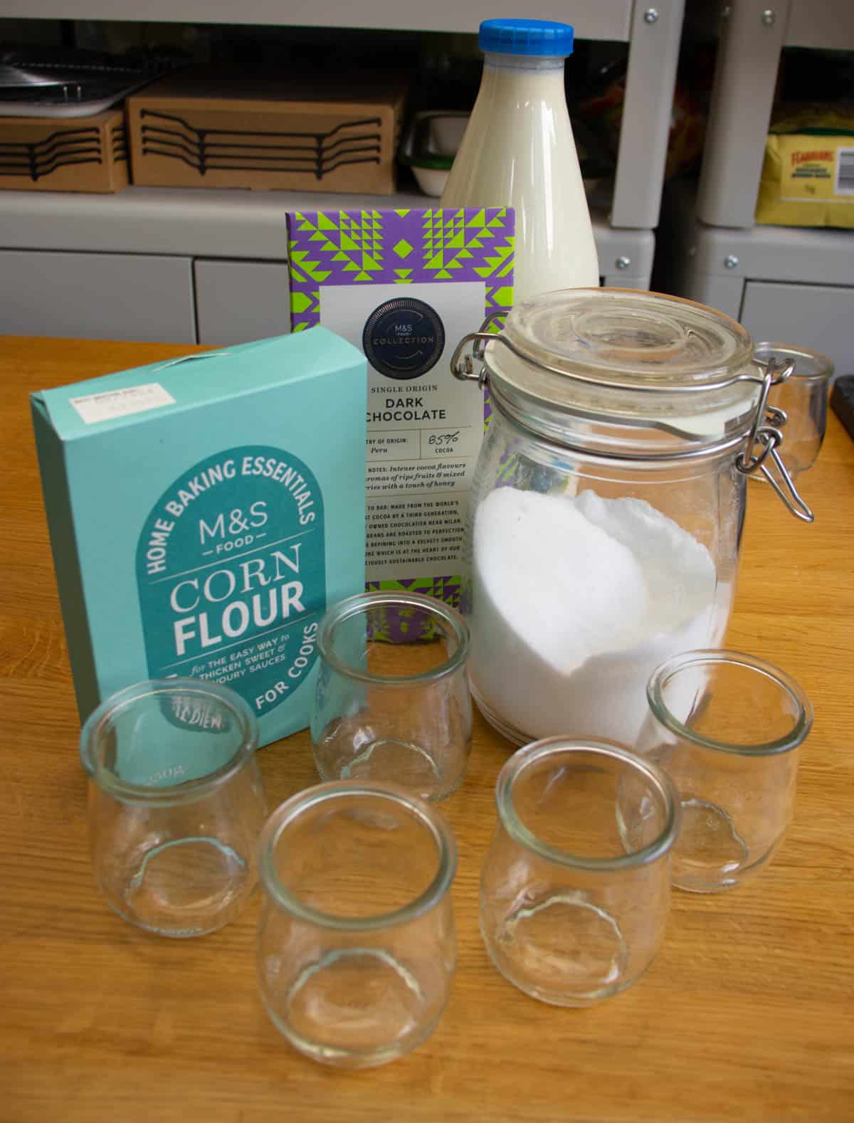 5 glass jars, packet of cornflour, kiln jar with sugar in it, bottle of milk and bar of chocolate all on wooden surface.
