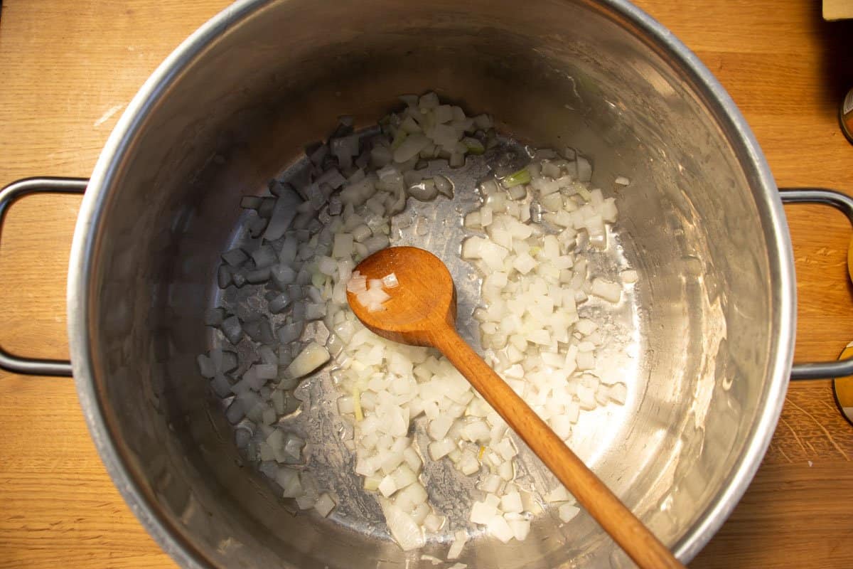 Chopped onion and oil in a saucepan on wooden surface being stirred by wooden spoon.