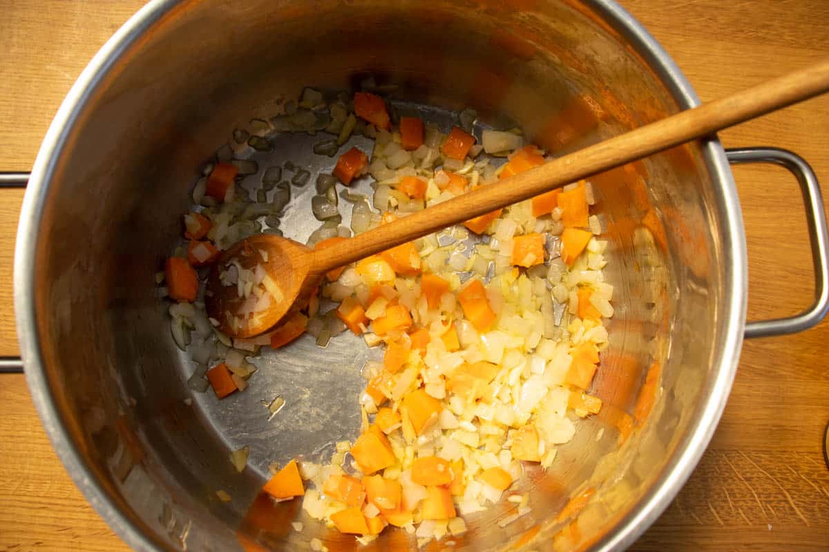 Chopped onion and carrots in a saucepan on wooden surface being stirred by wooden spoon.