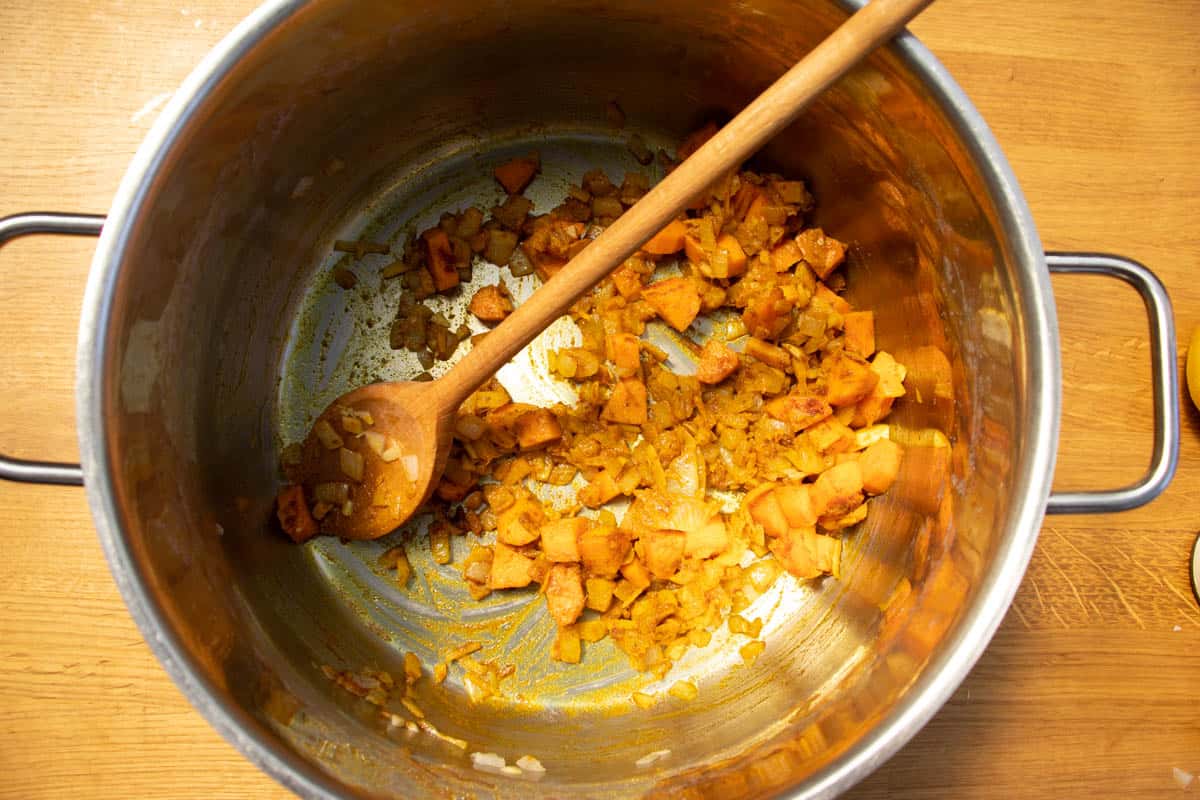 Chopped onion and carrots with ground spices in a saucepan on wooden surface being stirred by wooden spoon.