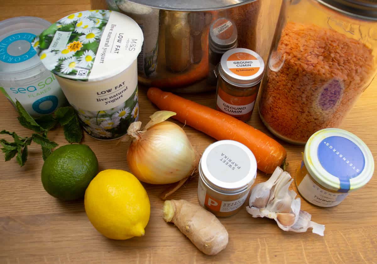 Ingredients for Mediterranean lentil soup on a wooden surface, lemon, ginger root, garlic, onion, jar of red lentils, tub of yoghurt, spice jars and tub of salt.