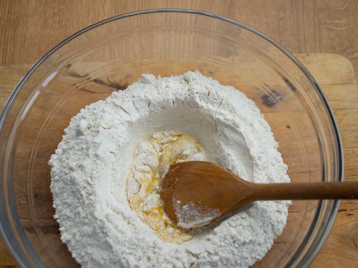 Egg being stirred into flour in glass mixing bowl with wood spoon.