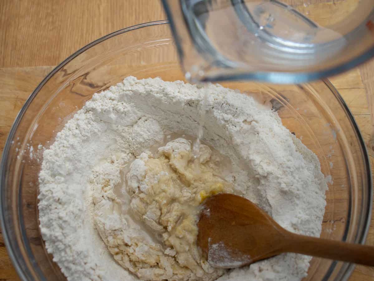 Water from jug being poured into flour and egg mixture in glass mixing bowl which is being stirred with wooden spoon.