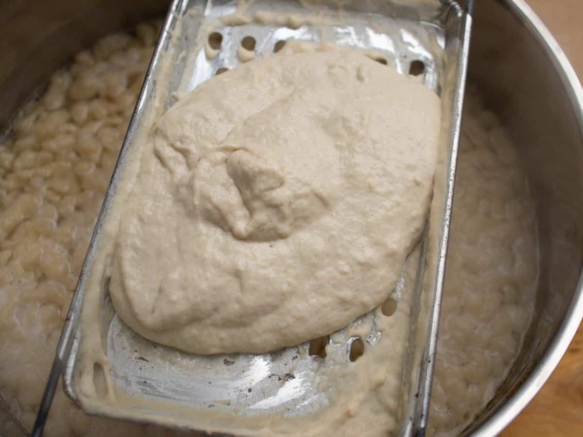Hungarian egg noodle dough mixture on Nokedli Szagatto held over saucepan of boiling water.
