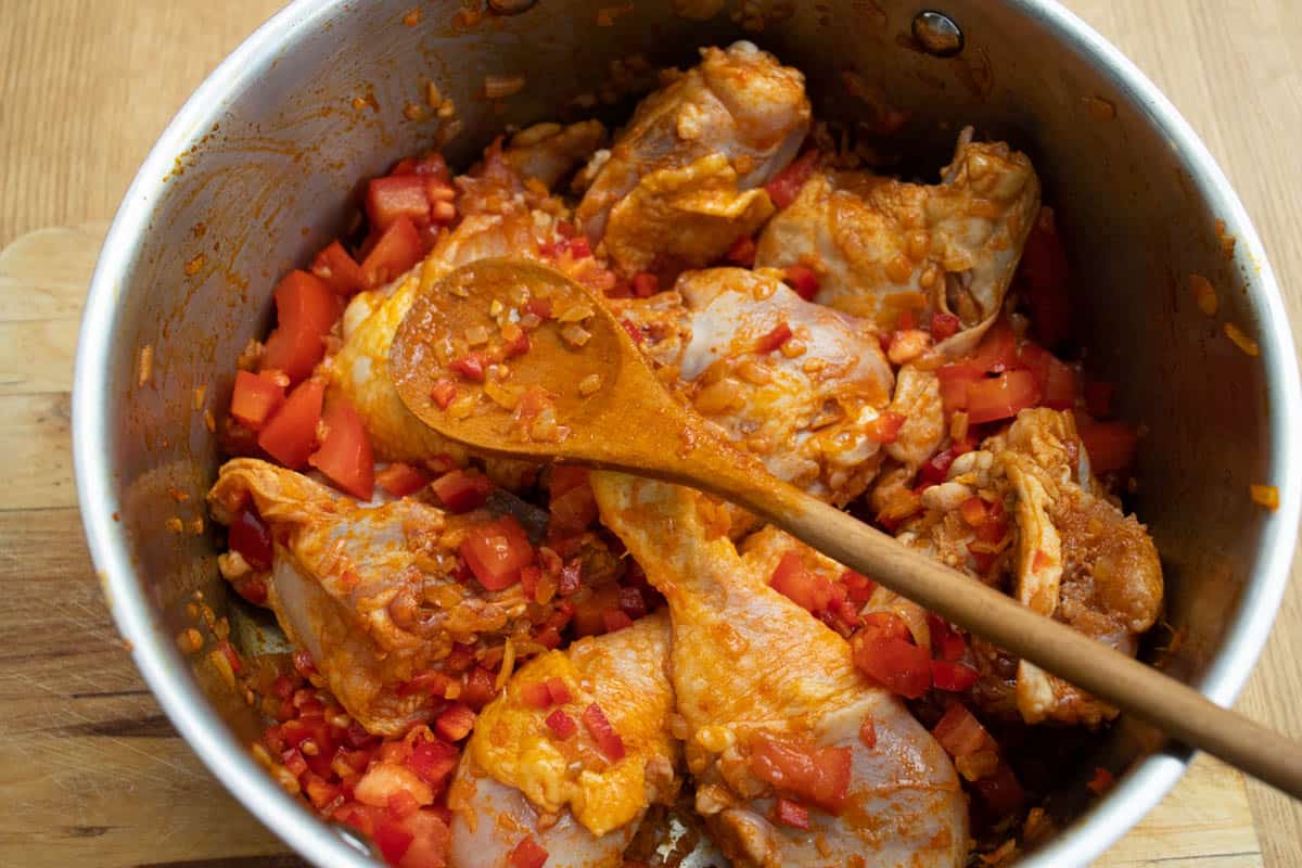 raw chicken pieces with chopped onion, peppers and tomatoes mixed with ground paprika spice in a saucepan with wooden spoon resting on mixture