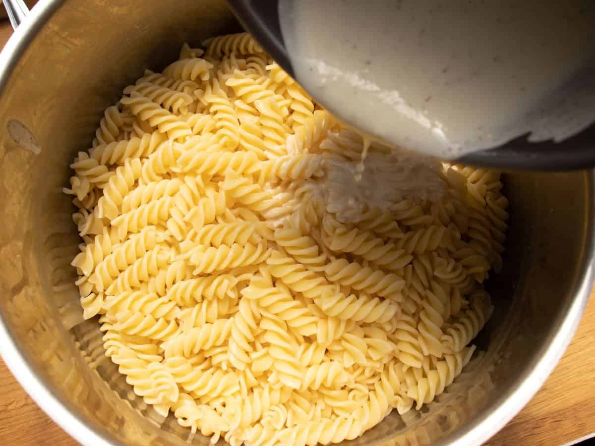 Cooked fusilli pasta in a saucepan with ladle of creme fraiche mixture being poured onto pasta.