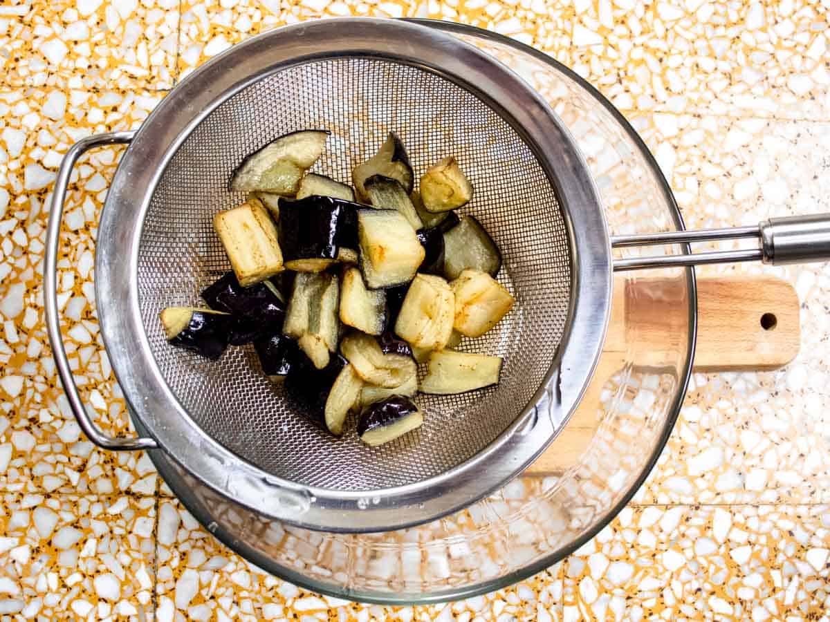 fried aubergine cubes in a stainless steel strainer held over a glass bowl