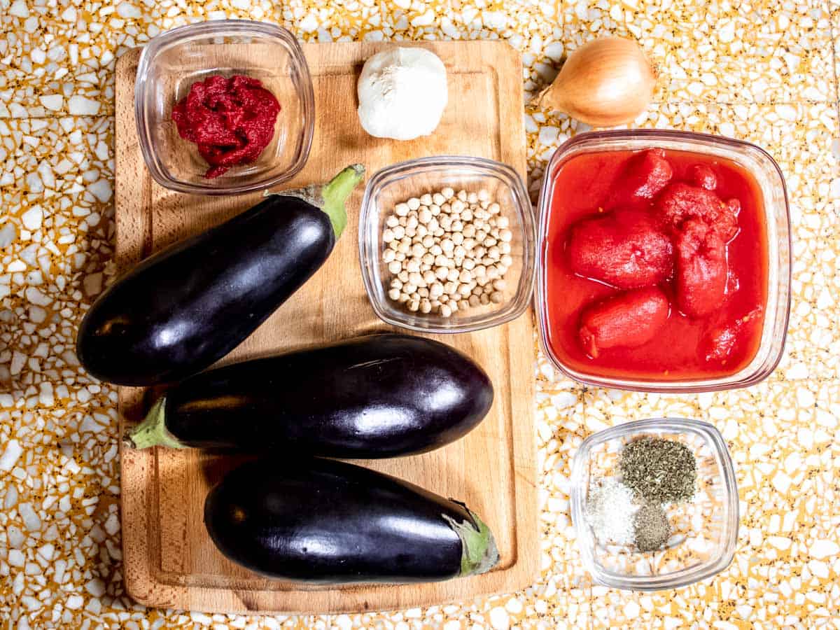 aubergines on a wooden board, chickpeas, spices, tinned tomatoes each in glass bowls, a garlic and an onion all laid out on marble background