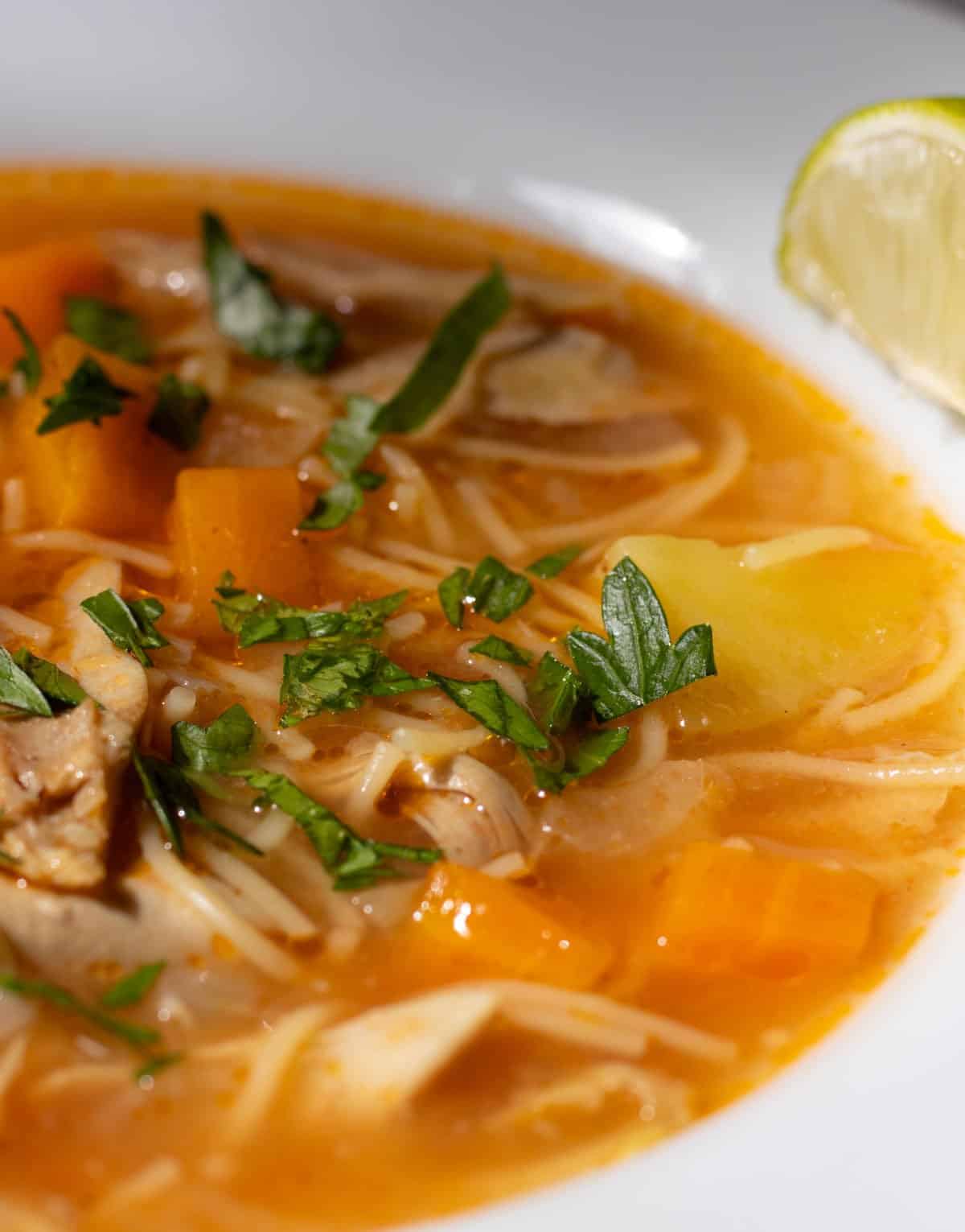 Close up of Cuban Chicken Noodle Soup sprinkled with parsley in serving bowl with slice of lime alongside.