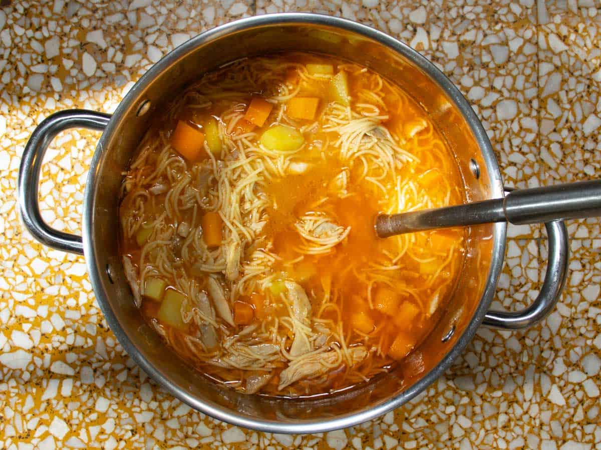 Cuban chicken noodle soup in stainless steel saucepan with stainless steel ladle.