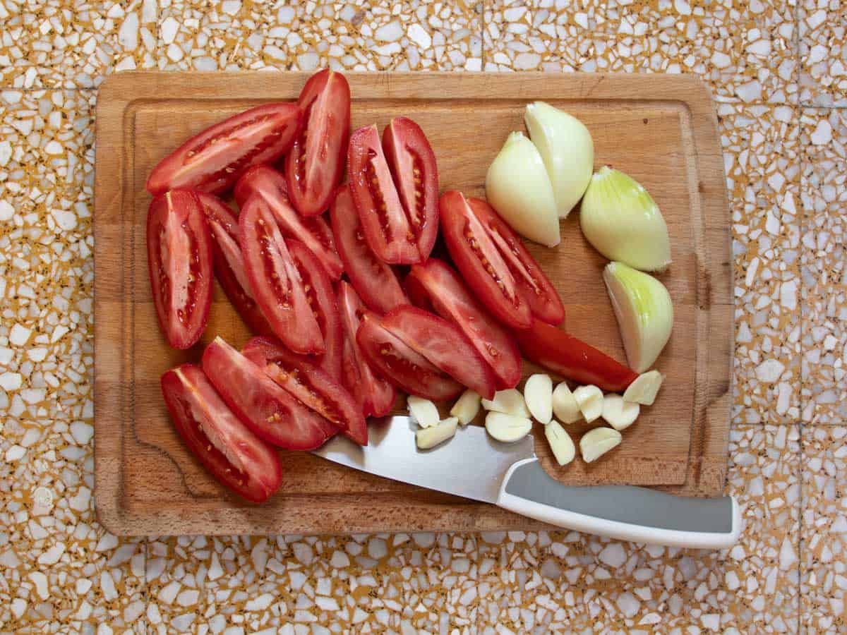 Sliced plum tomatoes, onions and garlic cloves with large sharp knife, all on wooden board.
