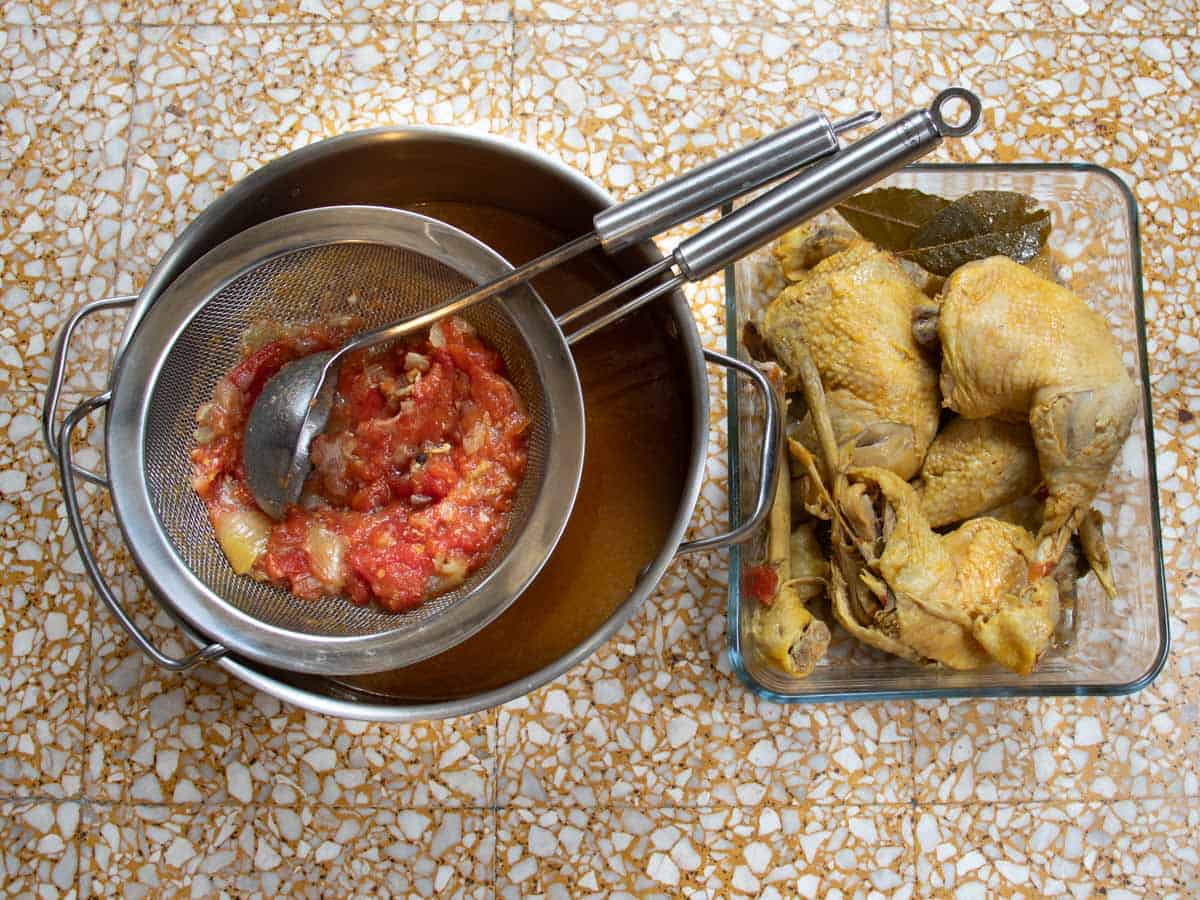 Cooked chicken pieces in a bowl alongside saucepan containing strained stock with strainer on top holding strained tomatoes and onions.