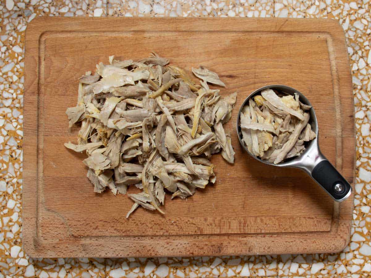 Shredded pieces of chicken on wooden chopping board with some in a stainless steel measuring cup.