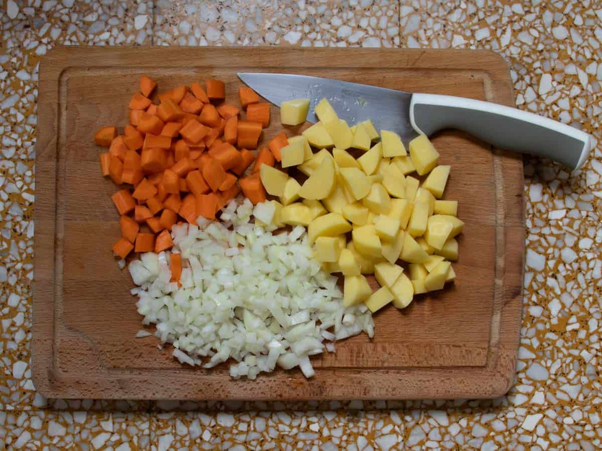 Diced carrots and potatoes, finely chopped onion with large sharp knife all on wooden chopping board.