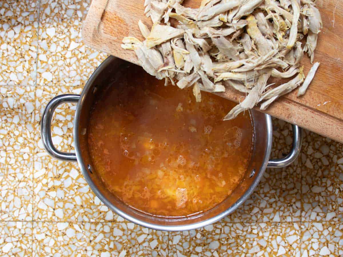 Shredded pieces of chicken being tipped into stainless steel saucepan containing Cuban chicken stock.
