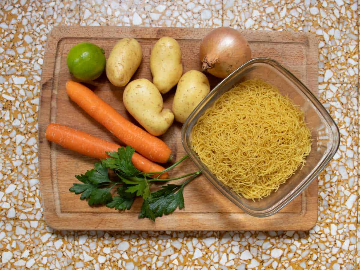 Potatoes, carrots, onion, lime, parsley, vermicelli in a glass bowl, all on a wooden board.
