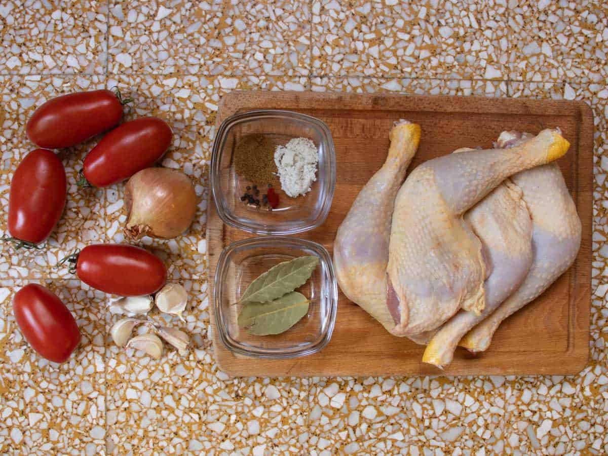 Plum tomatoes, onion, garlic cloves, bay leaves, salt, saffron, peppercorns, cumin and chicken pieces on a wooden board.