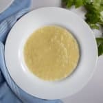 Celery and leek soup in a white bowl next to celery leaves and blue cloth.