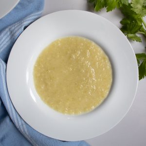 Celery and leek soup in a white bowl next to celery leaves and blue cloth.