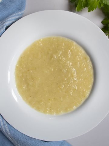 Celery and leek soup in a white bowl next to celery leaves and blue cloth.