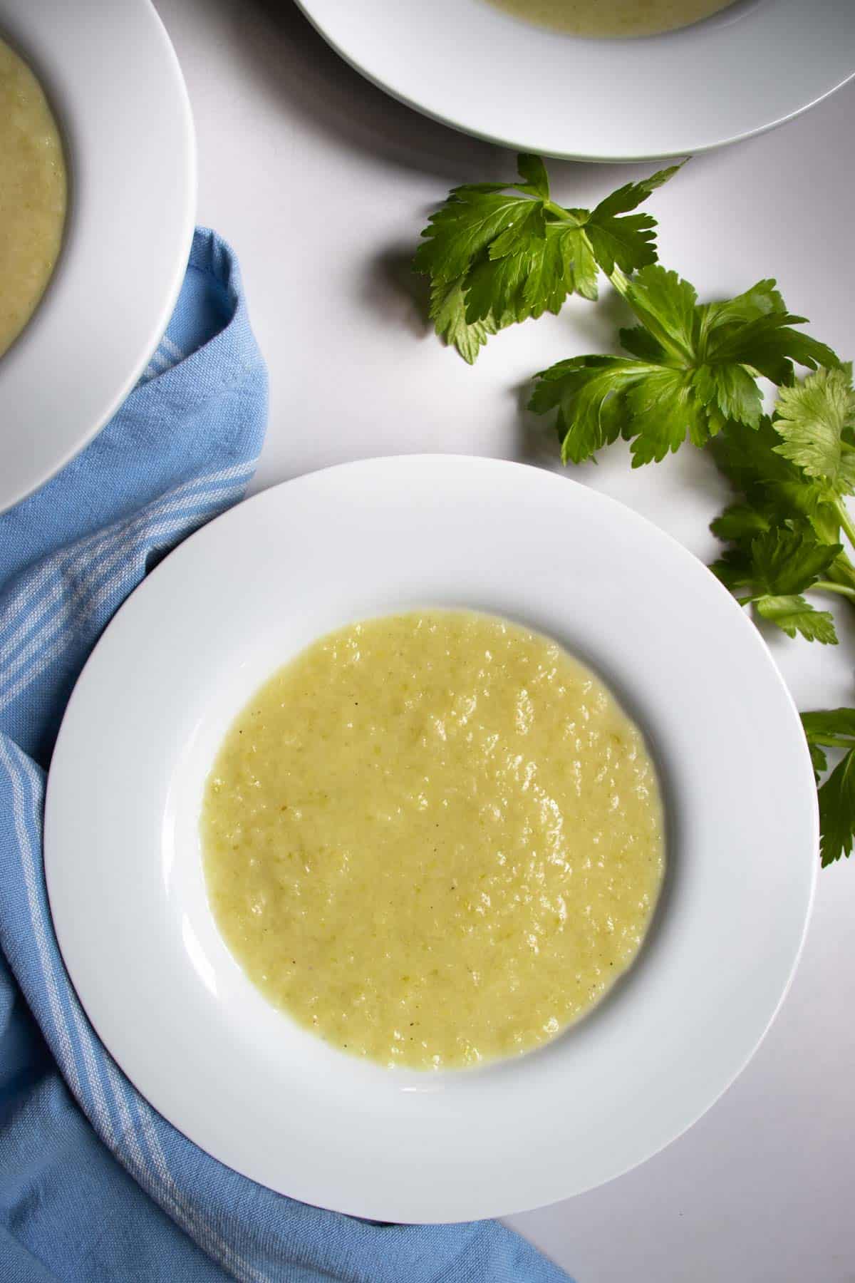 Celery and leek soup in white bowls next to celery leaves and blue cloth.