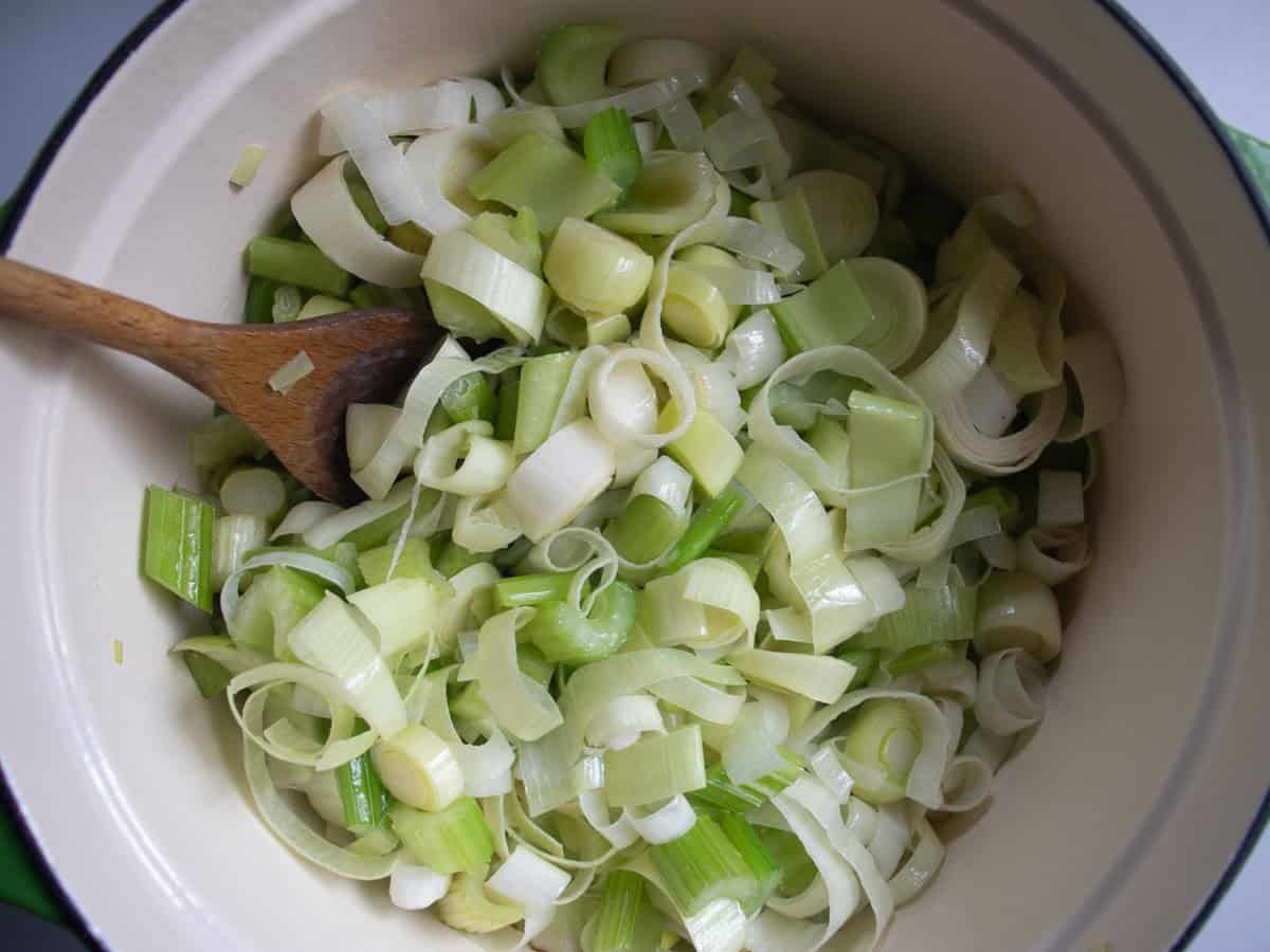 Chopped leeks and celery being stirred within wooden spoon in saucepan.