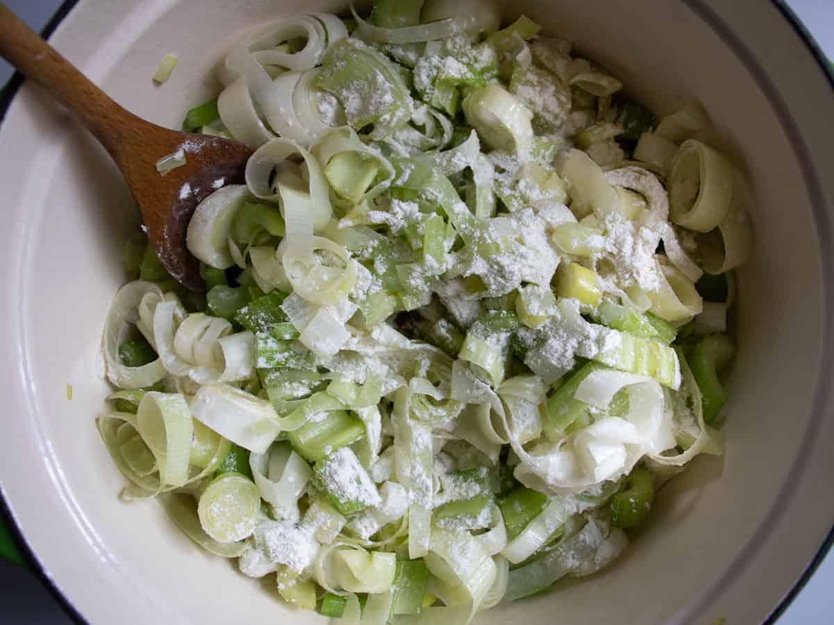 Chopped leeks and celery sprinkled with flour being stirred with wooden in saucepan.
