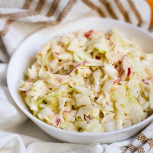 Apple coleslaw in white bowl on brown-striped cloth.