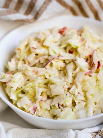 Apple coleslaw in white bowl on brown-striped cloth.