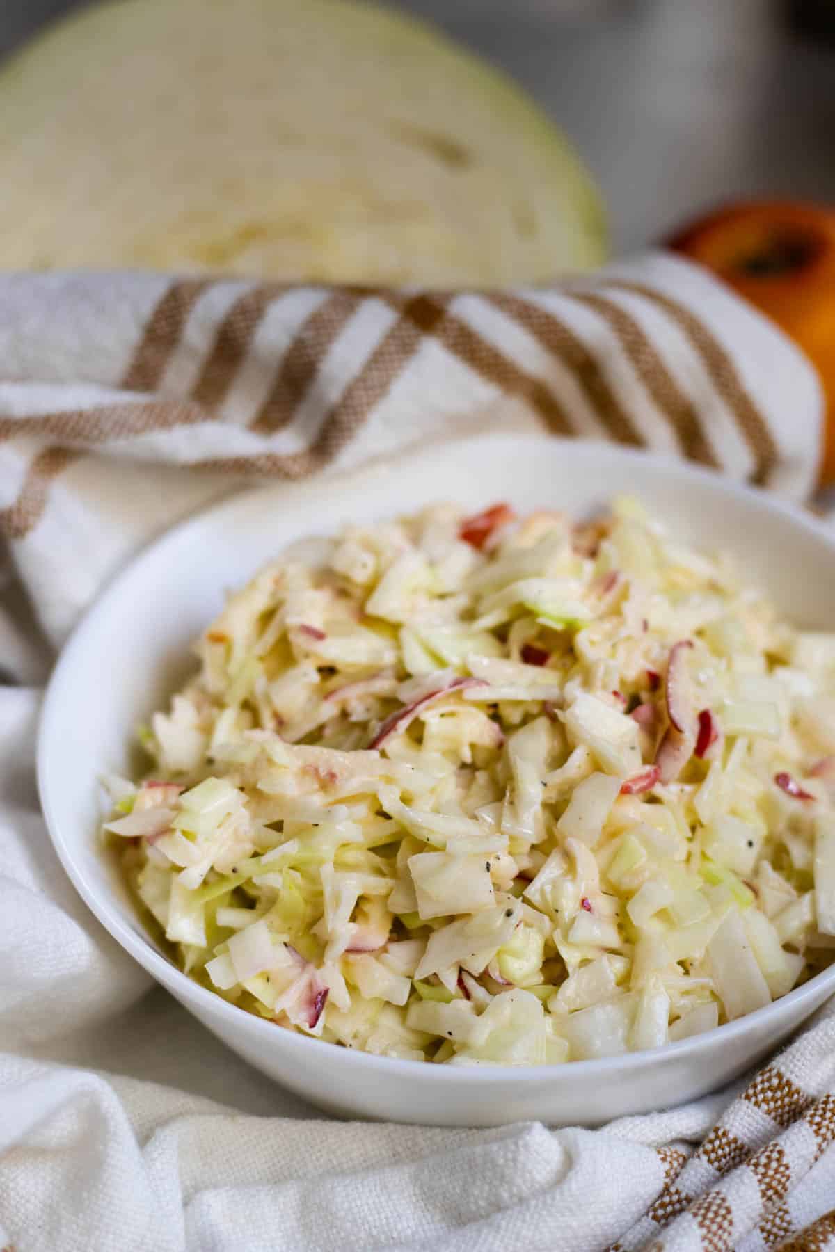 Apple coleslaw in white bowl on brown striped white cloth, with white cabbage and red apple in background.