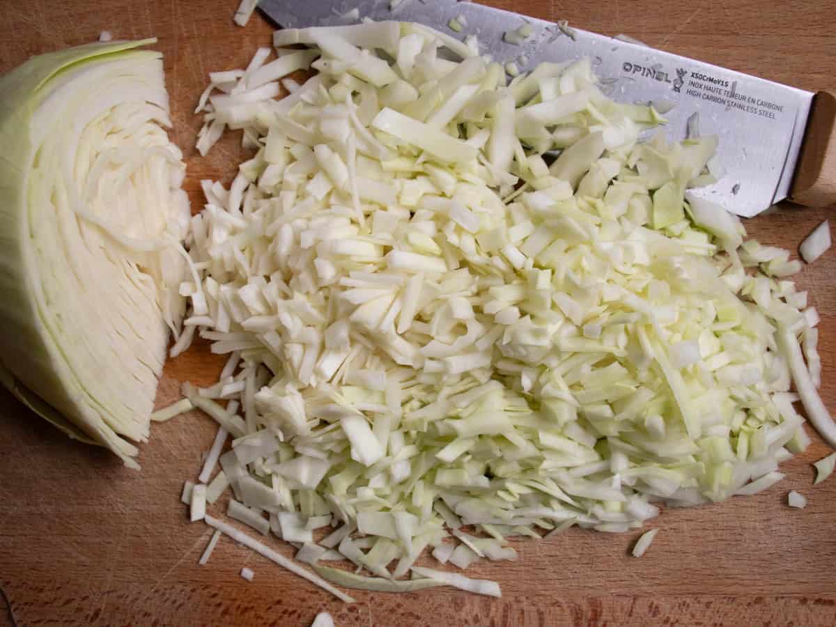 Section of white cabbage next to finely sliced cabbage and chef's knife on wooden chopping board.