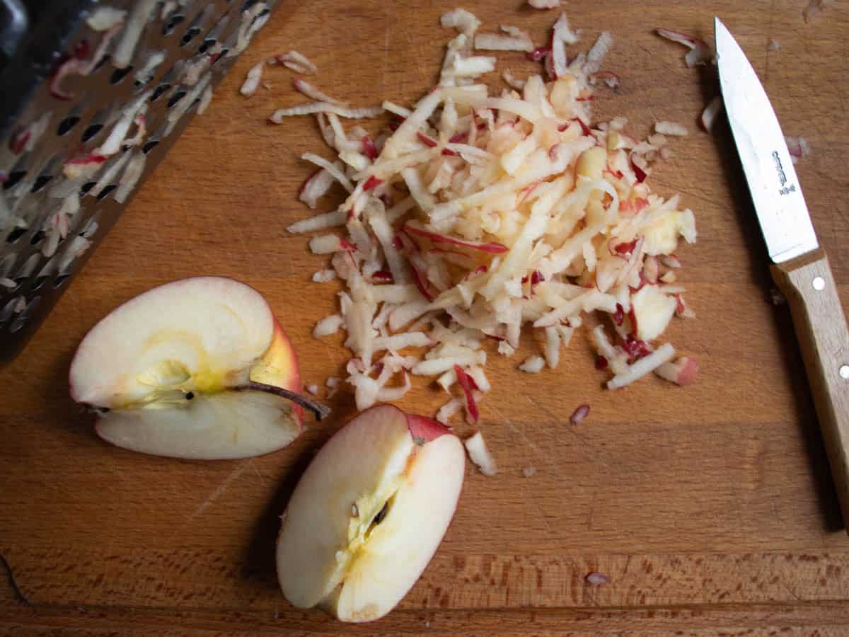 Quartered apple pieces next to grated apple and vegetable knife on wooden chopping board.