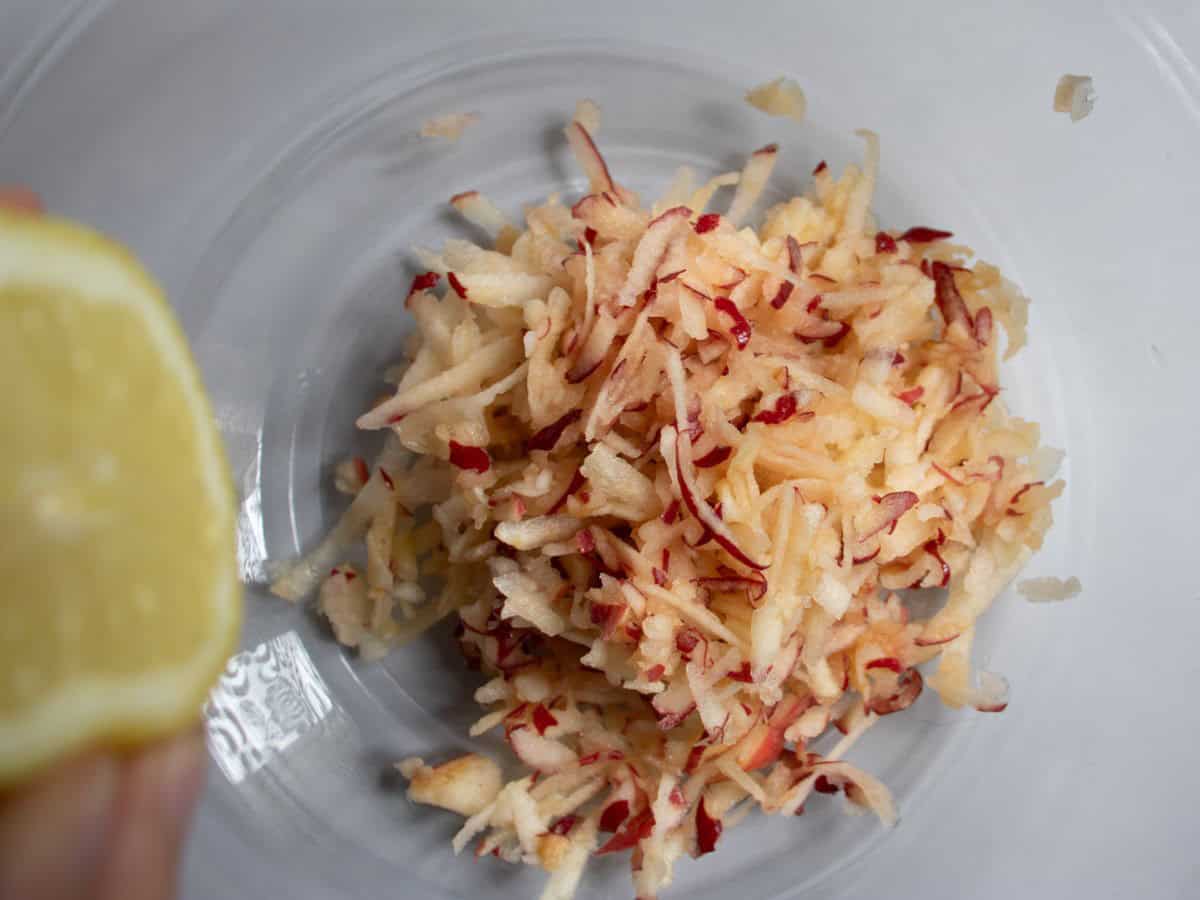 Lemon juice being squeezed onto grated apple in glass mixing bowl.