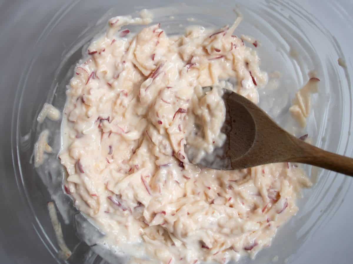 Grated apple, lemon juice and mayonnaise being stirred with wooden spoon in glass mixing bowl.