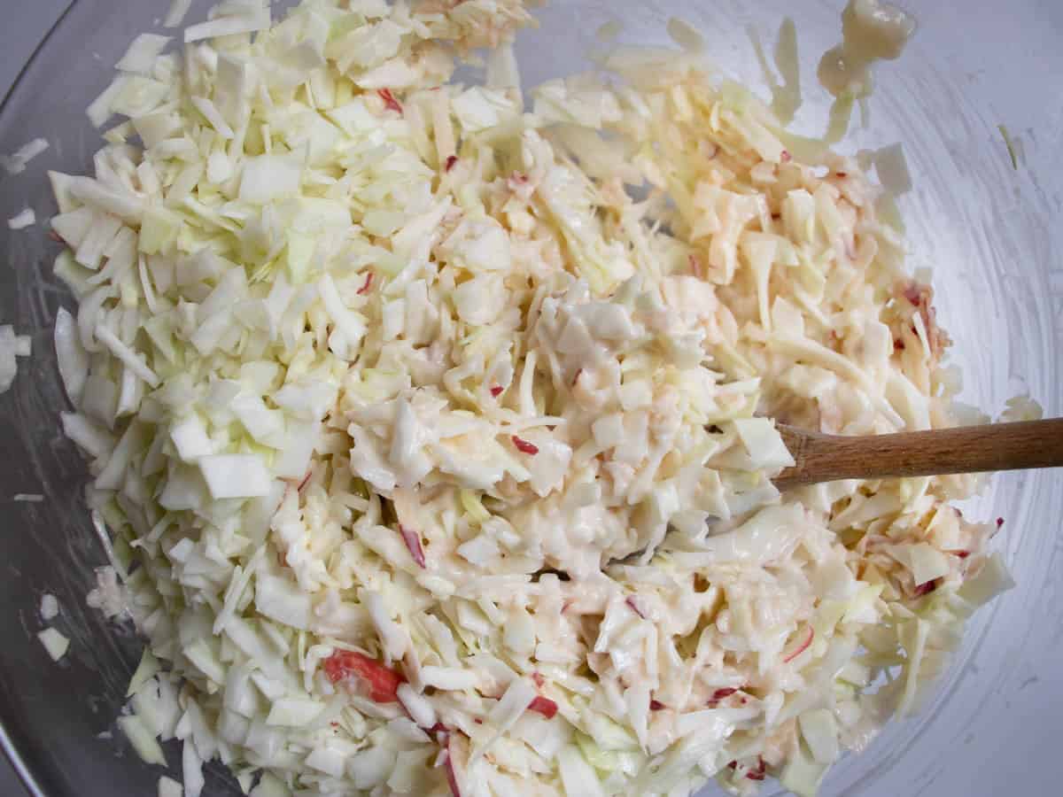 Chopped white cabbage, grated apple and mayonnaise being stirred in glass bowl with wooden spoon.