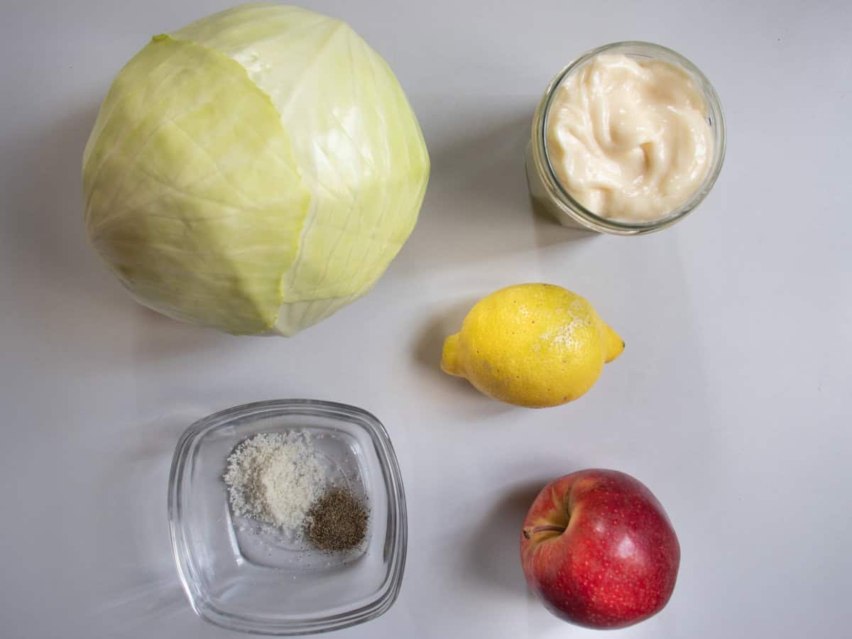 Ingredients for apple coleslaw laid out, white cabbage, mayonnaise, lemon, red apple, salt and pepper.