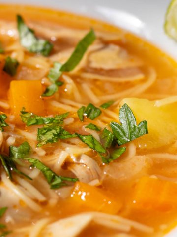 Cuban chicken noodle soup in a bowl sprinkled with chopped parsley and with a slice of lime on the side.