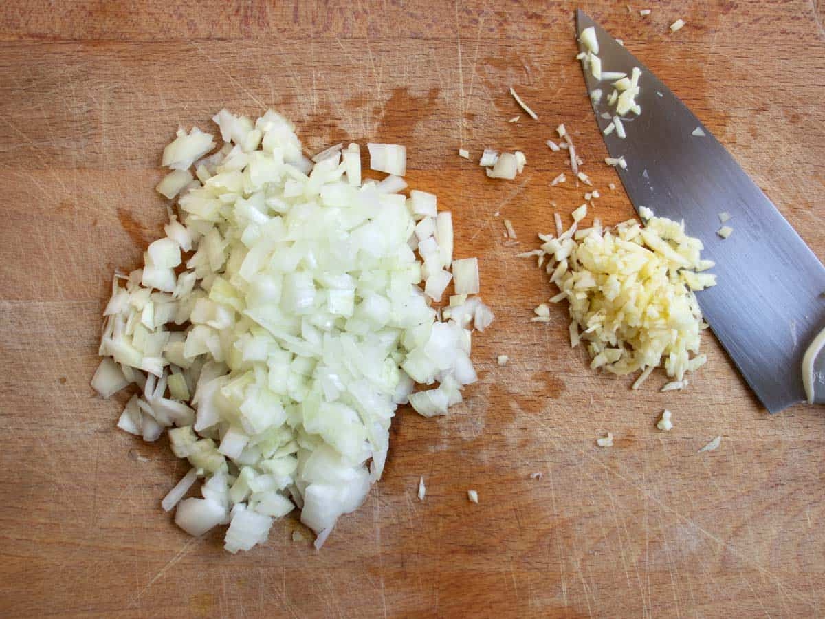 Finely chopped onion and garlic on wooden chopping board with knife alongside.
